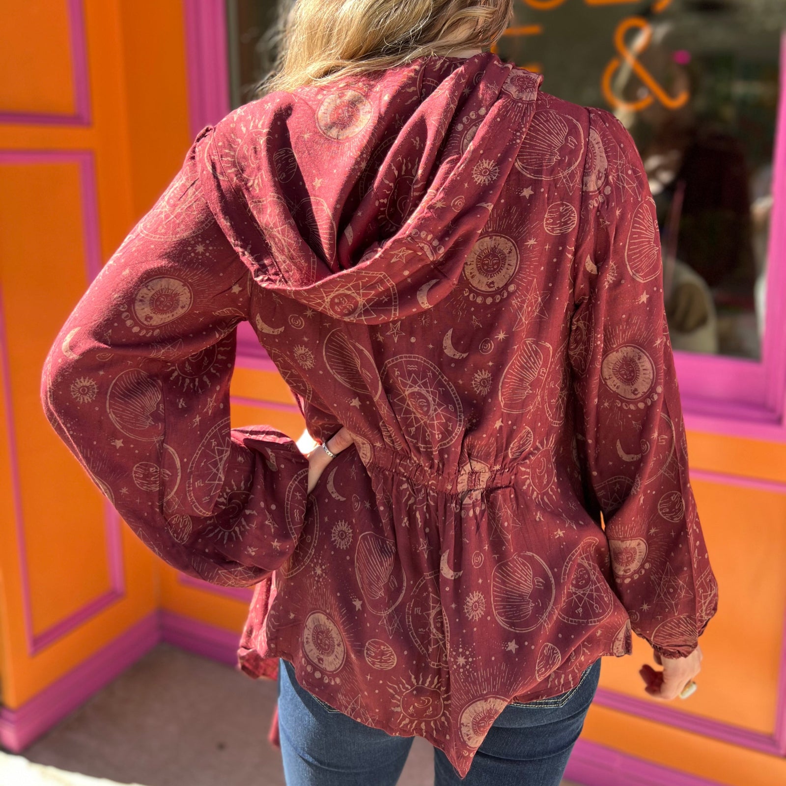 A woman wearing the Flowy Hoodie Waterfall Hooded Cardigan with a black tank and bell bottoms in front of a pink and orange wall.