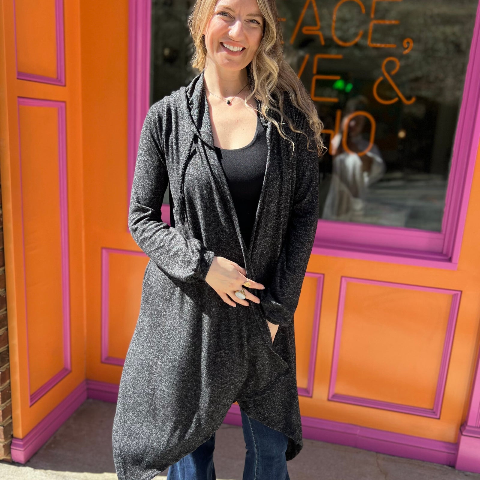 A woman wearing the black Moon Cycle Waterfall Hoodie with a black tank and bellbottoms in front of an orange and pink wall.
