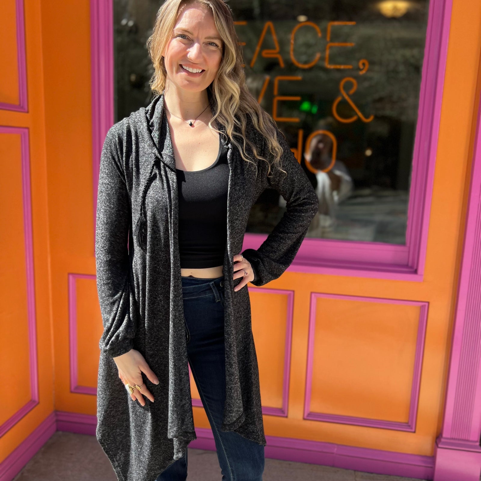 A woman wearing the black Moon Cycle Waterfall Hoodie with a black tank and bellbottoms in front of an orange and pink wall.
