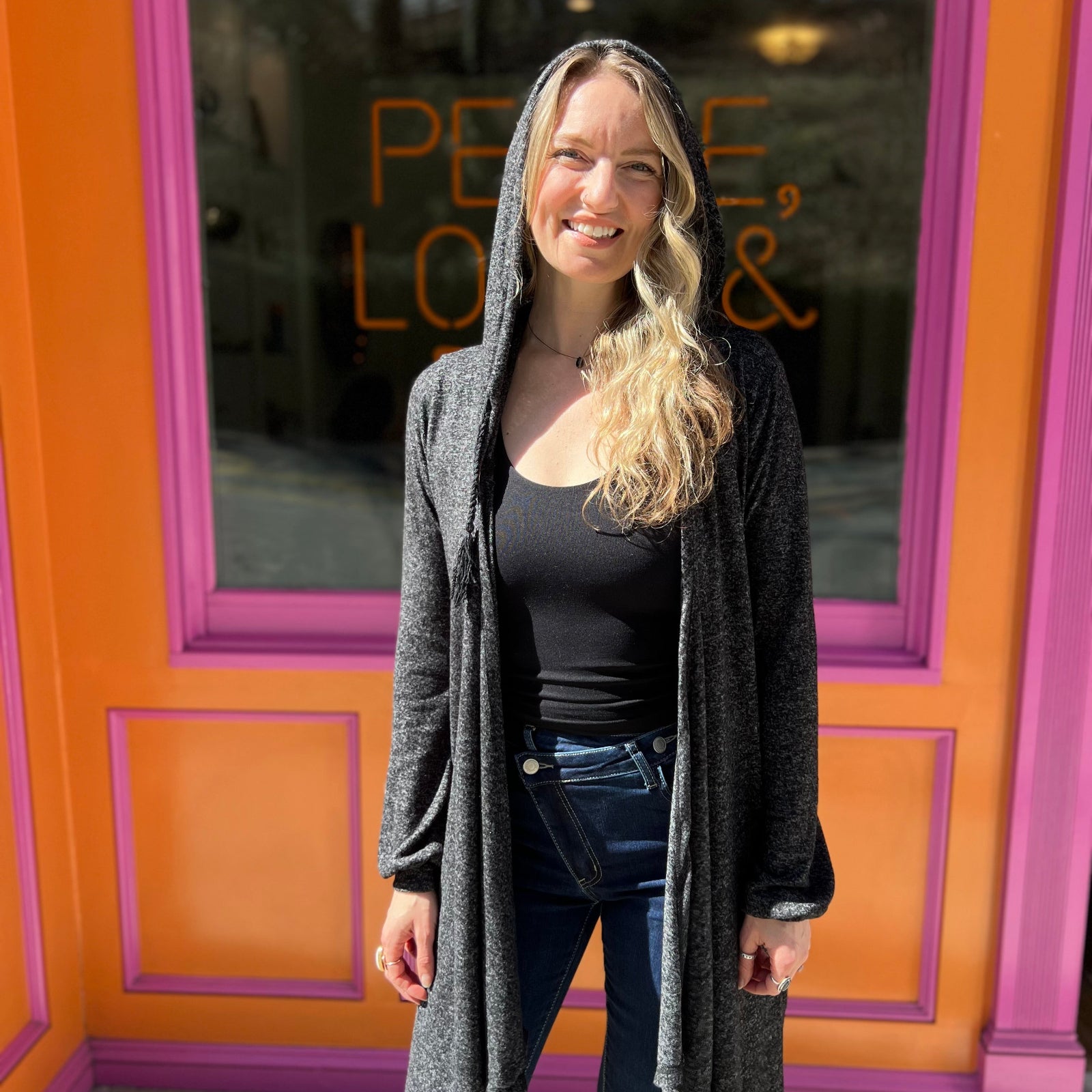A woman wearing the black Moon Cycle Waterfall Hoodie with a black tank and bellbottoms in front of an orange and pink wall.
