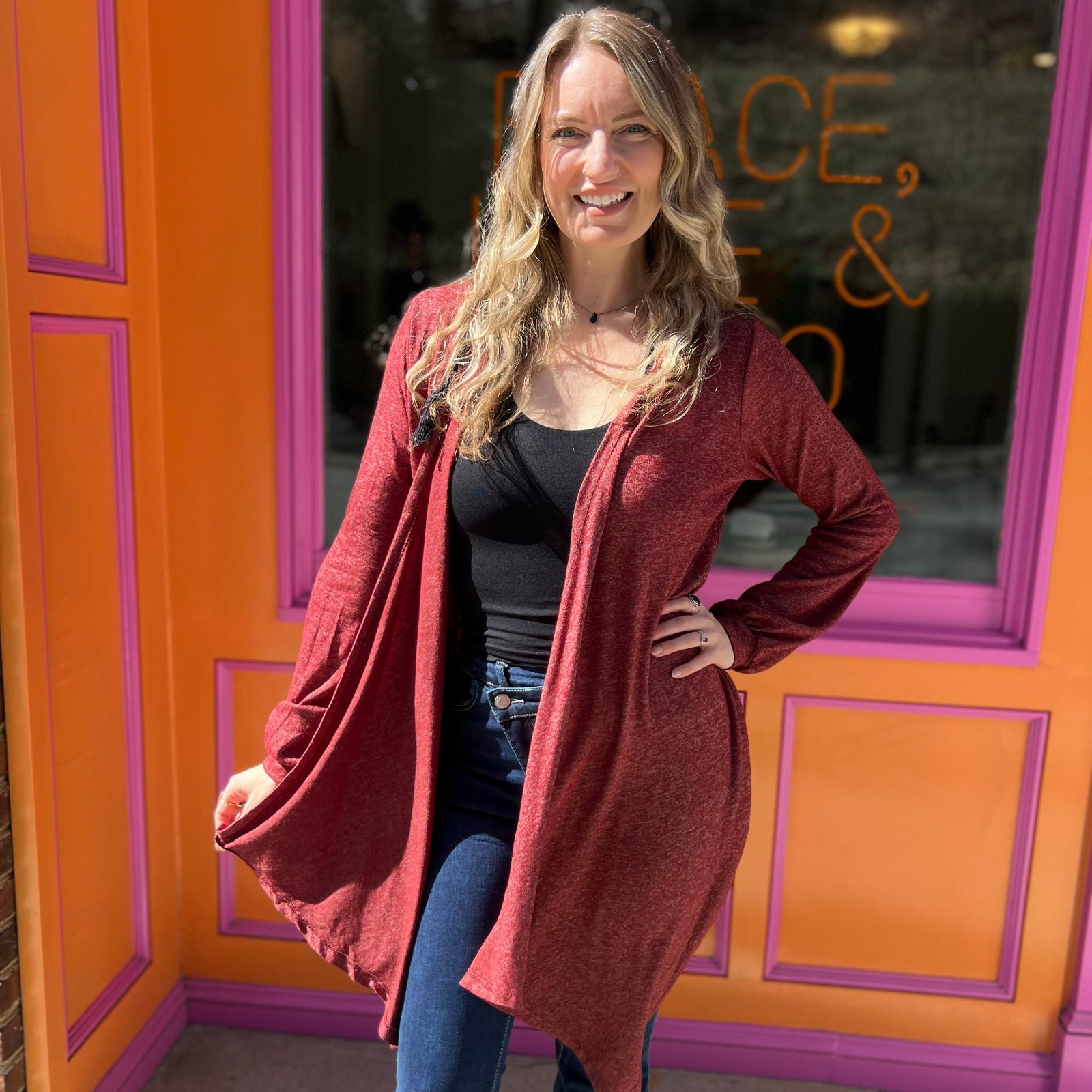 A woman wearing the maroon Moon Cycle Waterfall Hoodie with a black tank and bellbottoms in front of an orange and pink wall.