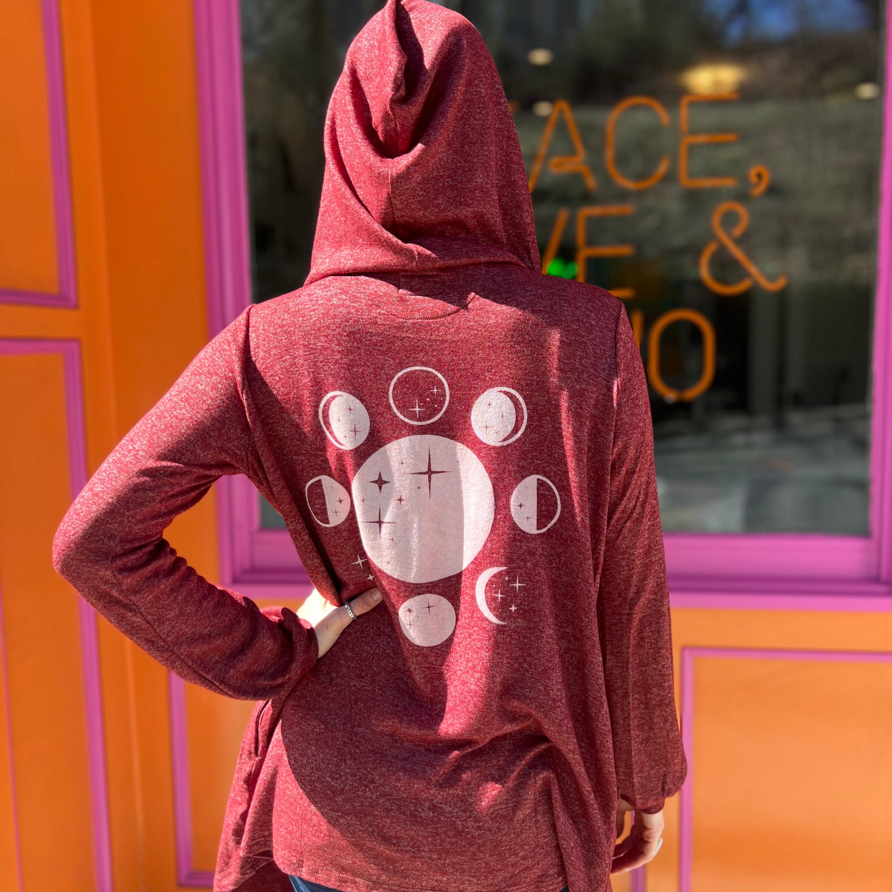 A woman wearing the maroon Moon Cycle Waterfall Hoodie with a black tank and bellbottoms in front of an orange and pink wall.