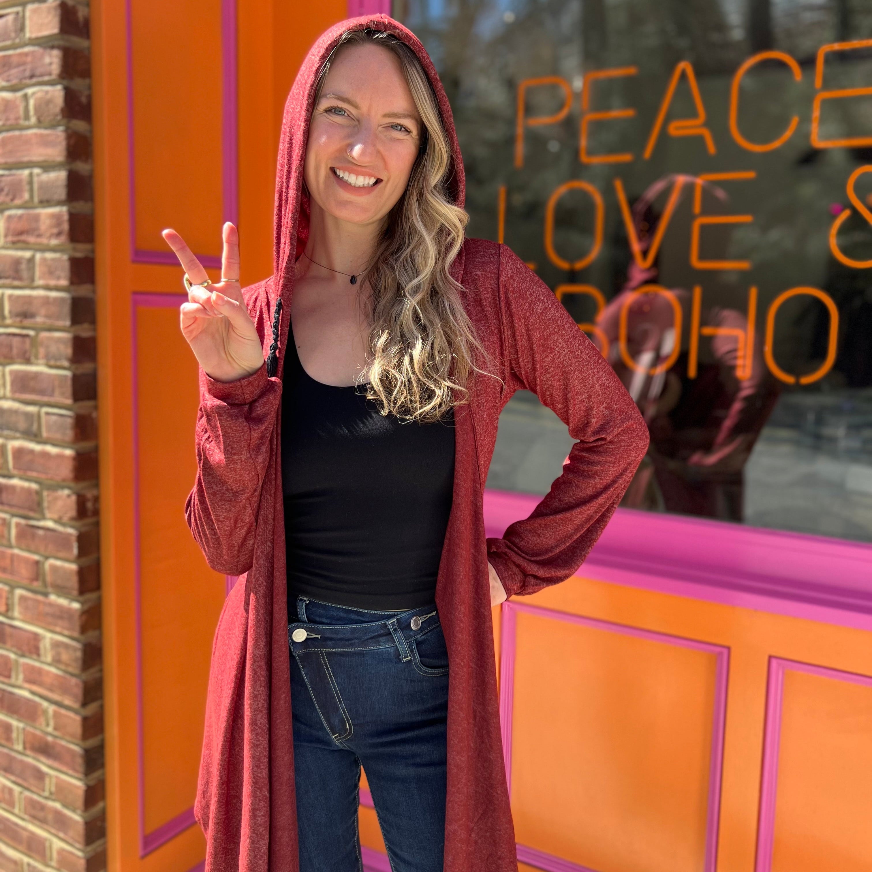 A woman wearing the maroon Moon Cycle Waterfall Hoodie with a black tank and bellbottoms in front of an orange and pink wall.