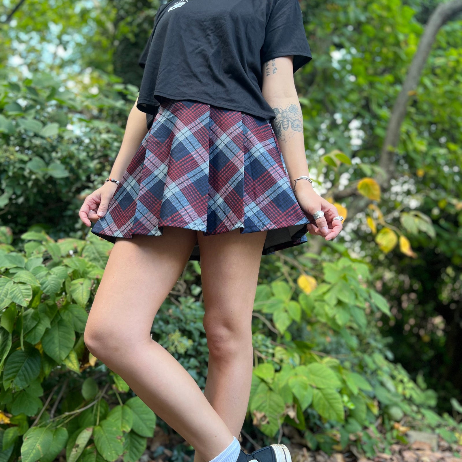 A woman wearing the Plaid Pleated Mini Skirt with a black shirt in front of greenery.