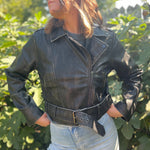 A woman wearing the Washed Black Leather Moto Jacket with jeans in front of some greenery.