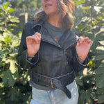 A woman wearing the Washed Black Leather Moto Jacket with jeans in front of some greenery.