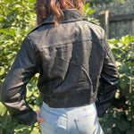 A woman wearing the Washed Black Leather Moto Jacket with jeans in front of some greenery.