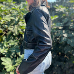 A woman wearing the Washed Black Leather Moto Jacket with jeans in front of some greenery.