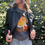 A woman wearing the Washed Black Leather Moto Jacket with jeans in front of some greenery.