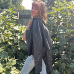 A woman wearing the Washed Black Leather Moto Jacket with jeans in front of some greenery.