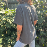 A woman wearing the Season Of The Witch Halloween Retro Cotton T-Shirt with jeans in front of some greenery, showing off the side.