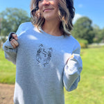 A woman wearing the Halloween Skeletons Gray Cotton Crewneck Sweatshirt and holding the embroidered graphic out.