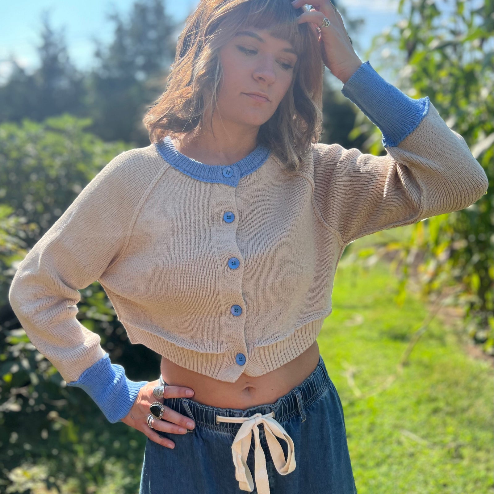A woman wearing the Contrast Knitted Crew Neck Button Down Cardigan Sweater with denim pants in front of greenery.