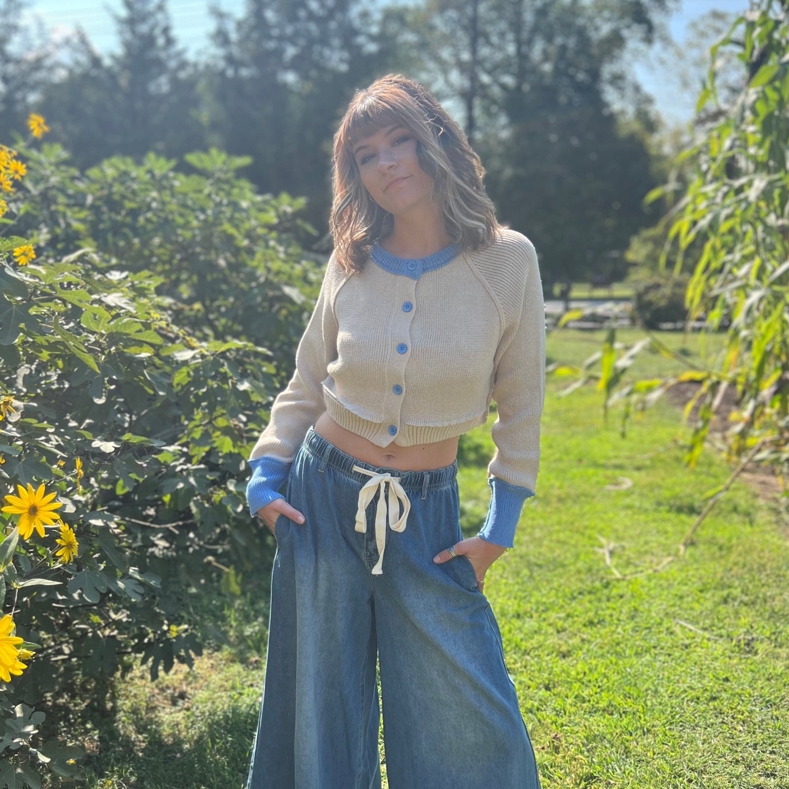 A woman wearing the Contrast Knitted Crew Neck Button Down Cardigan Sweater with denim pants in front of greenery.