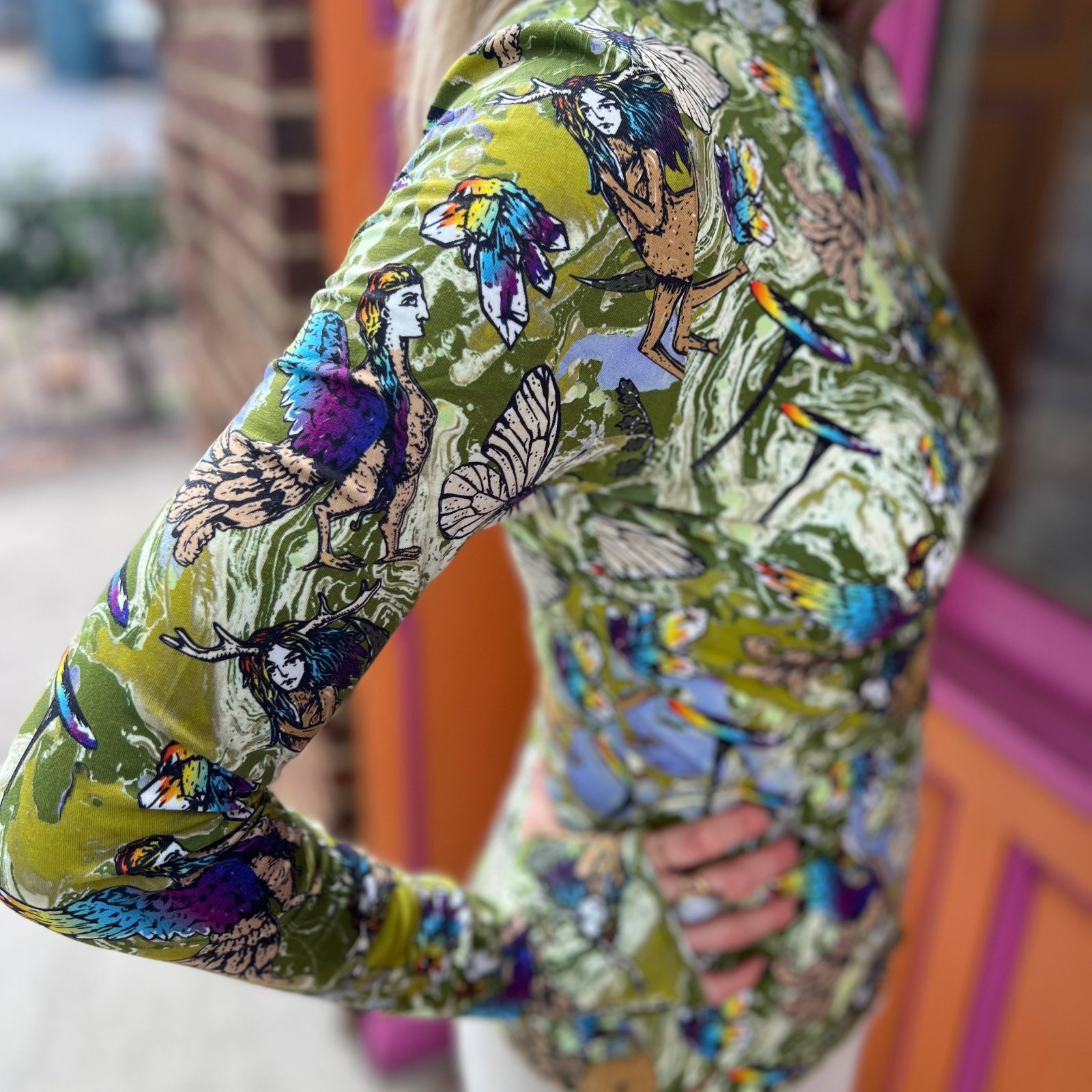 A woman wearing the green Cosmic Mythology Long Sleeve Top with beige linen pants in front of a window.