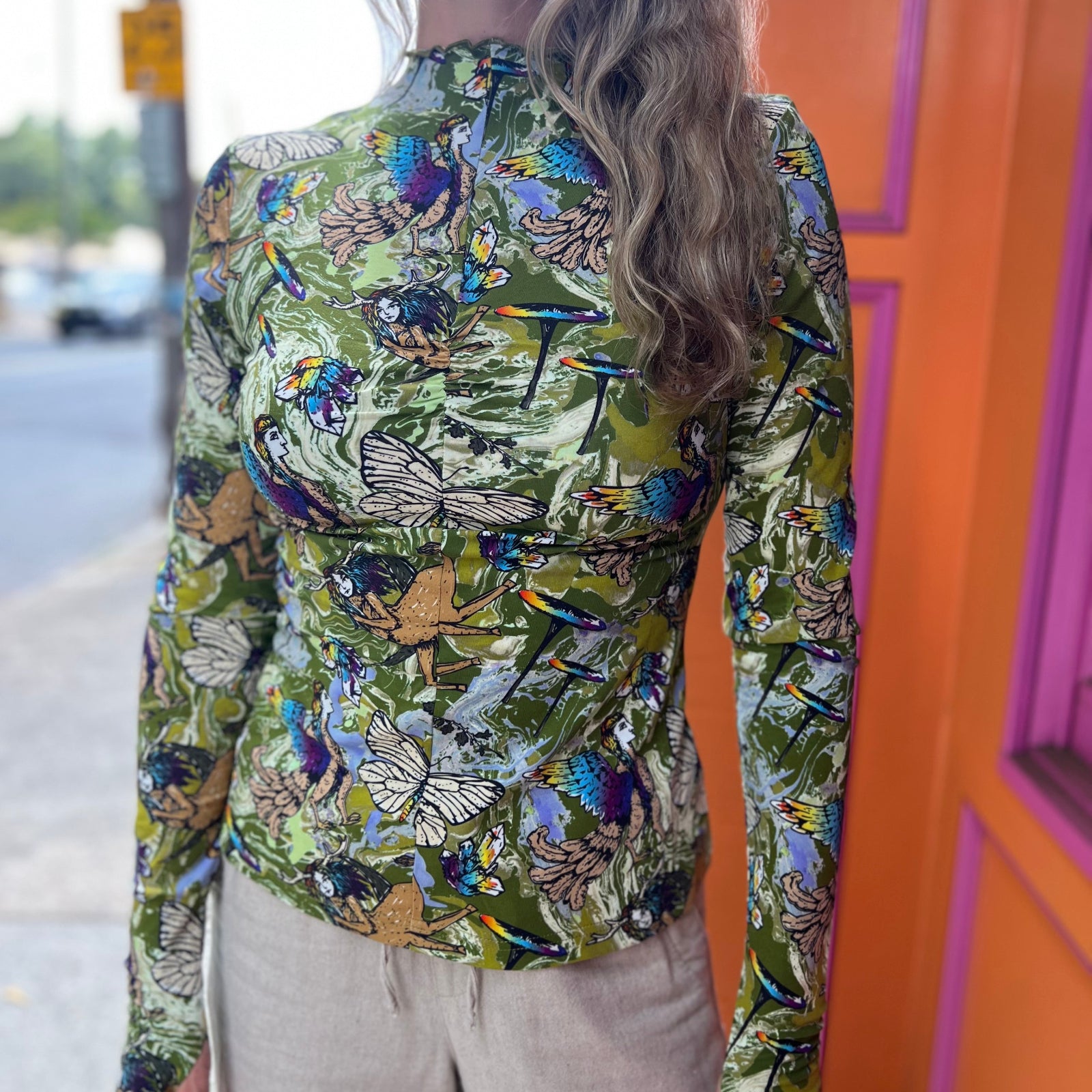 A woman wearing the green Cosmic Mythology Long Sleeve Top with beige linen pants in front of a pink and orange wall.