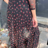 Back close up view of a woman wearing a black mesh cherry print maxi dress, showing the sheer detail and attached slip.