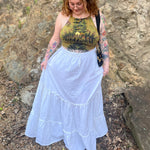 A woman wearing a tie dye top with the white Homespun Tiered Maxi Skirt in front of a rock wall.