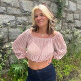 Woman wearing a blush pink crochet knit crop top and dark jeans posing confidently in front of a stone wall with greenery