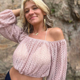Woman with two-tone hair and off-shoulder pink top smiling with one arm up, standing against a rock wall