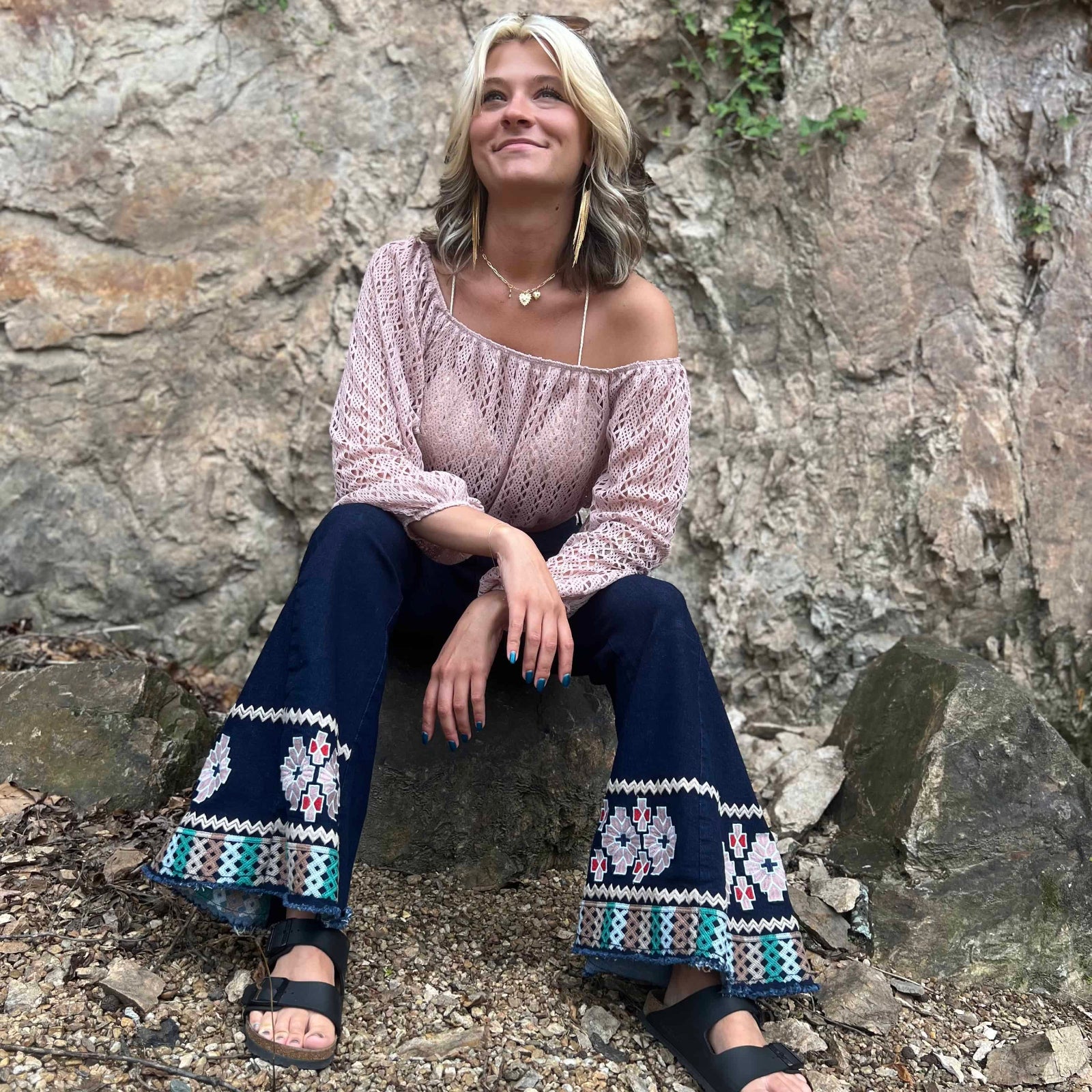 Woman sitting on a rock in embroidered flare jeans and pink crochet crop top, smiling and looking upward
