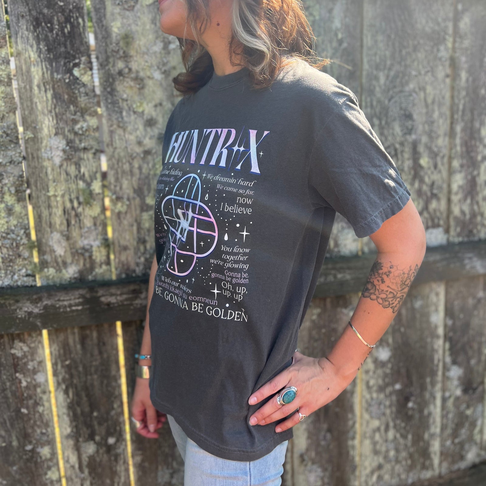 A woman wearing the Huntrix Kpop Demon Hunters Inspired Cotton Tee with jeans in front of a wooden fence, showing off the side.
