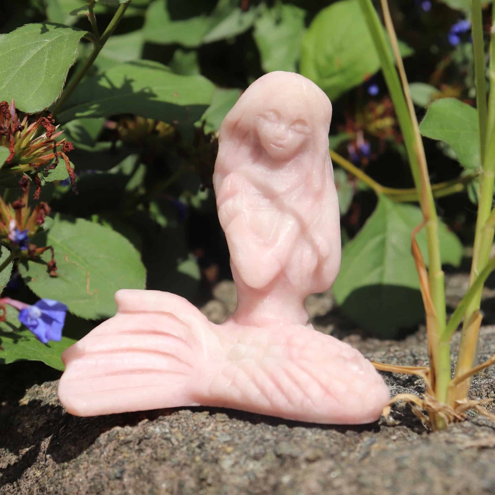 Pink opal crystal mermaid sculpture of a reclining figure on a natural background with greenery and flowers.