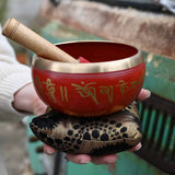 Red singing bowl with gold text held by a person in front of a rustic background