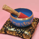 Blue singing bowl with gold accents and a wooden mallet on a decorative cloth against a pink background