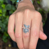 Hand wearing a silver ring with a decorative design against a blurred green background