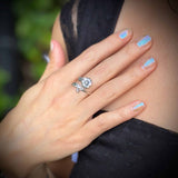 Hand wearing a silver ring with a flower design, against a blurred natural background.