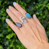 Hand wearing three rings with gemstones against a green leafy background