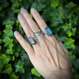 Hand wearing multiple rings with gemstones against a green leafy background