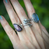 Hand wearing multiple silver rings with purple stones against a blurred green background