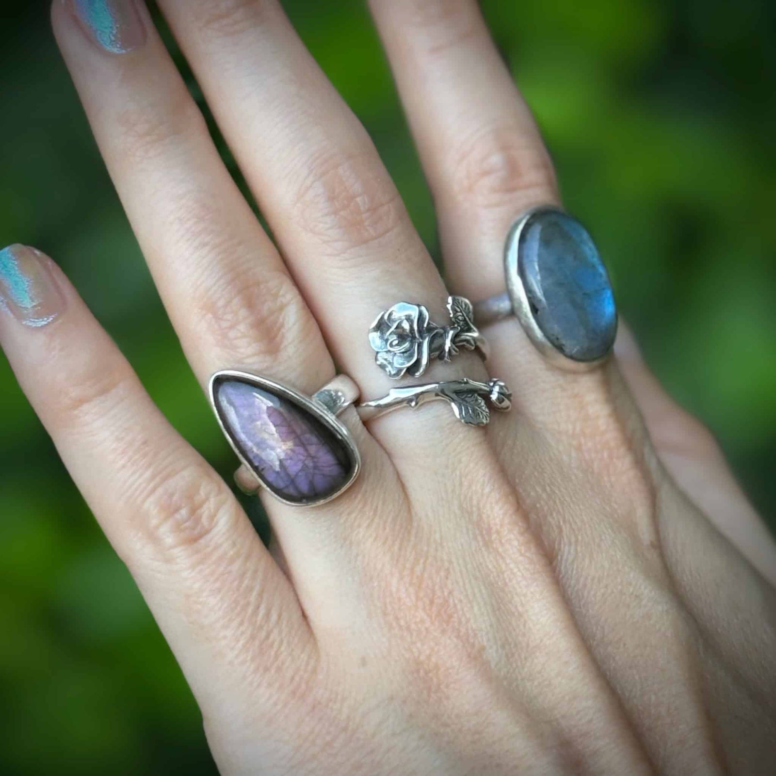 Hand wearing multiple silver rings with purple stones against a blurred green background