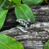 Silver ring with a leaf design on a wooden log surrounded by green leaves