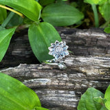 Silver ring with a floral design on a wooden log surrounded by green leaves