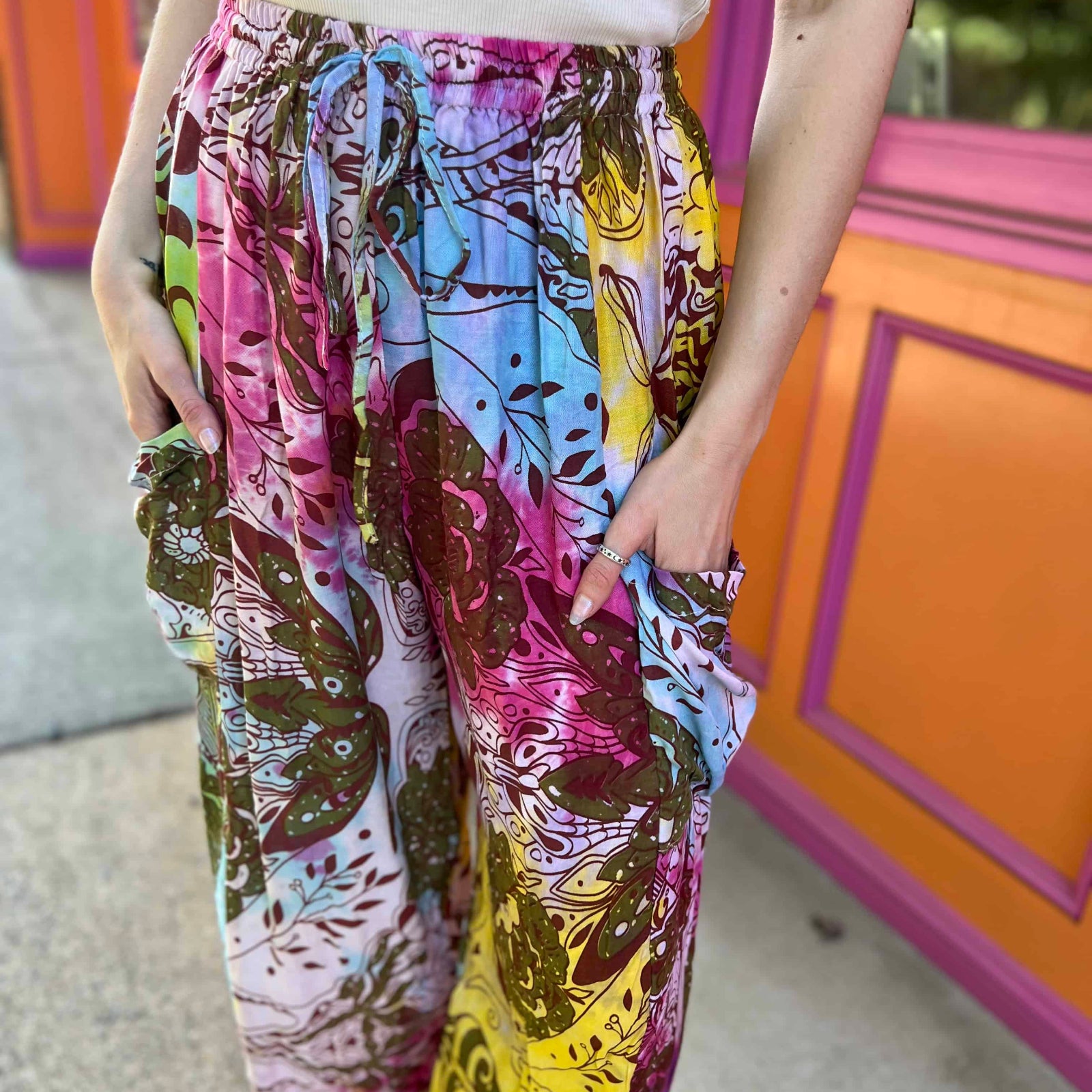Rainbow tie-dye cargo pants with green floral moth pattern with both hands in pockets and close up of draw string