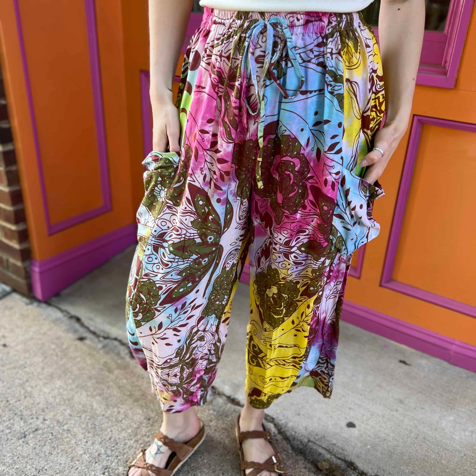 Rainbow tie-dye cargo pants with green floral moth pattern close up view with both hands in pockets