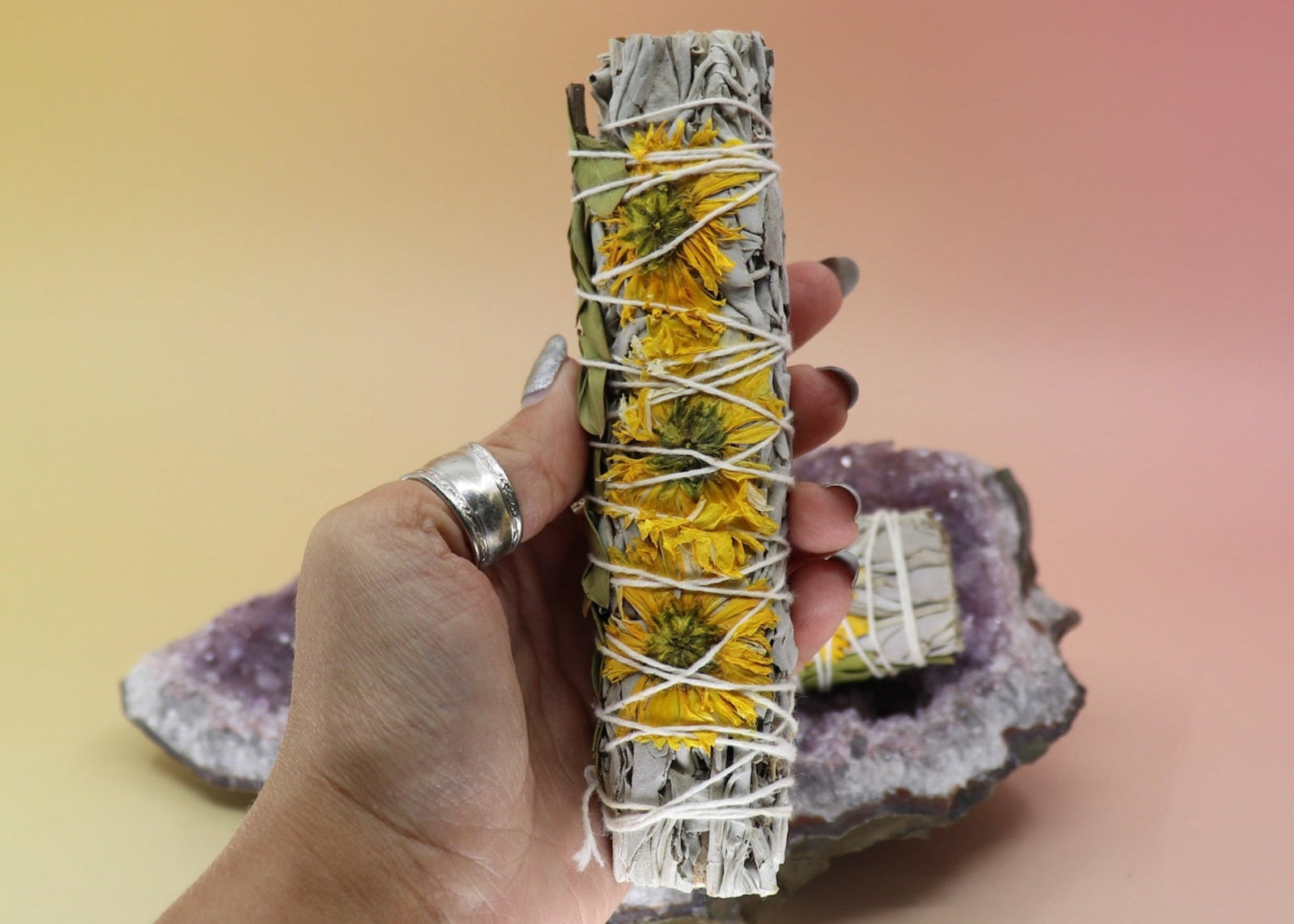 Someone holding the Organic Sunflower White Sage - 4 in. Smudge Stick in front of an amethyst piece all on a pink and yellow background.