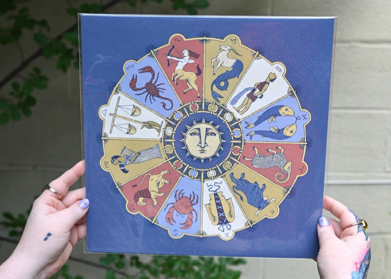 The Mystical Zodiac Print being held in front of a cream-colored wall with greenery.