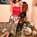 A woman wearing the Sequin Flare Thunderbolt Disco Pants with a pink fringe sequin top while sitting down in a velvet chair.