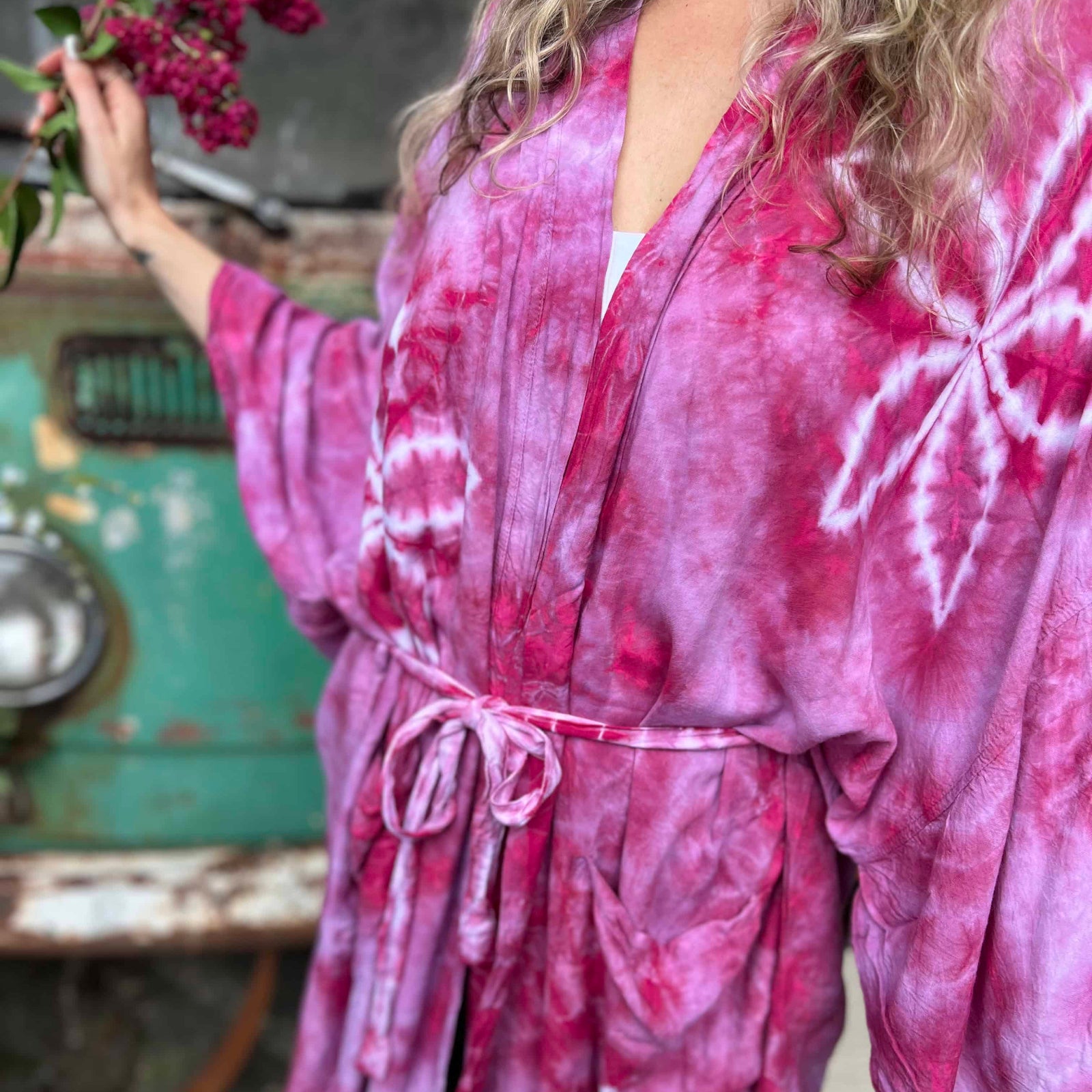 Person wearing a pink tie-dye robe holding flowers in an outdoor setting.