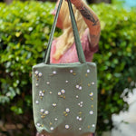 Blonde woman wearing a floral dress and carrying a sage green linen tote bag with hand-embroidered white daisies