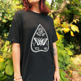 A woman wearing the Dump Him Ouija Cotton T-Shirt with a navy and red plaid skirt in front of greenery.