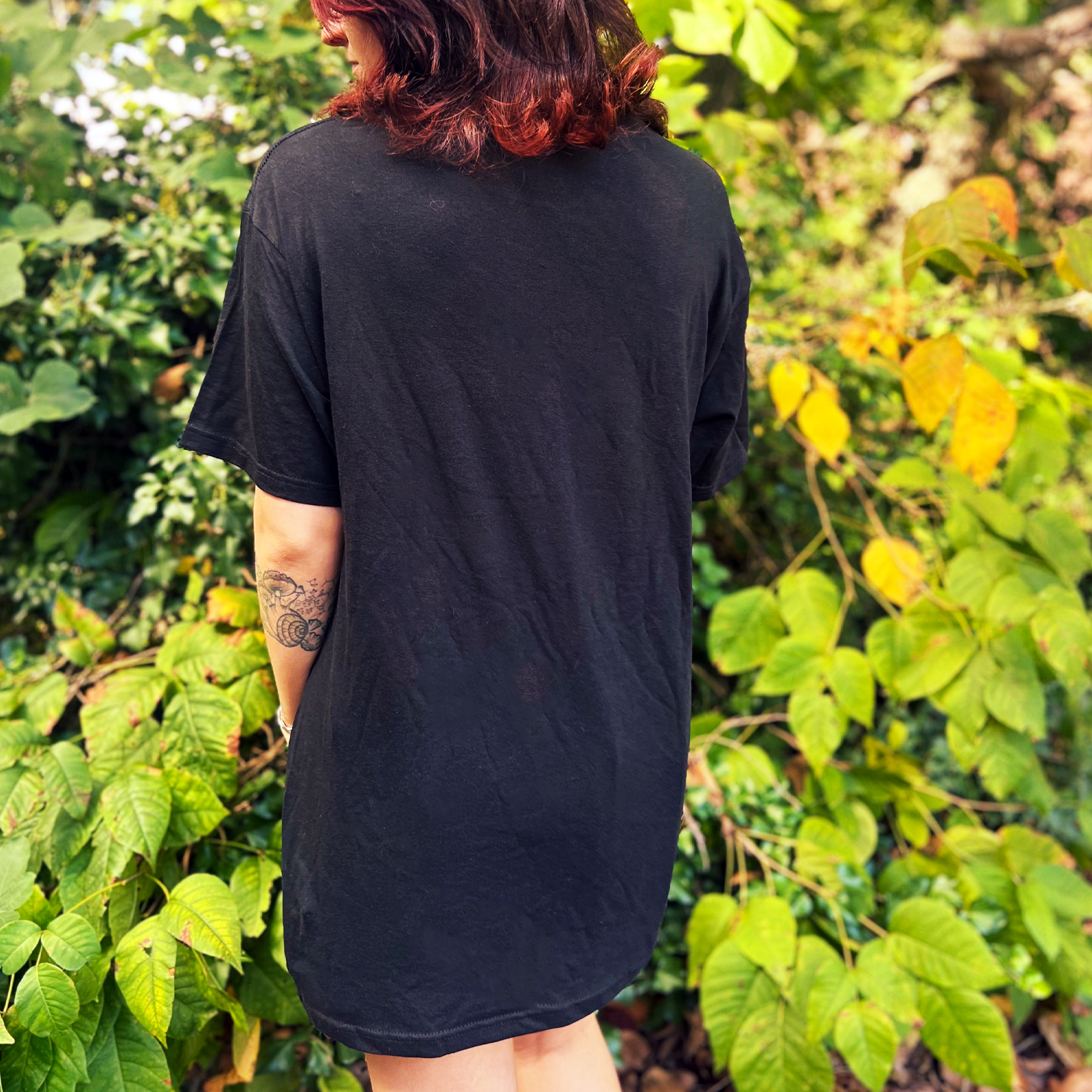 A woman wearing the Dump Him Ouija Cotton T-Shirt with a navy and red plaid skirt in front of greenery, showing off the back.