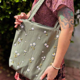 Blonde woman wearing a floral dress and carrying a sage green linen tote bag with hand-embroidered white daisies