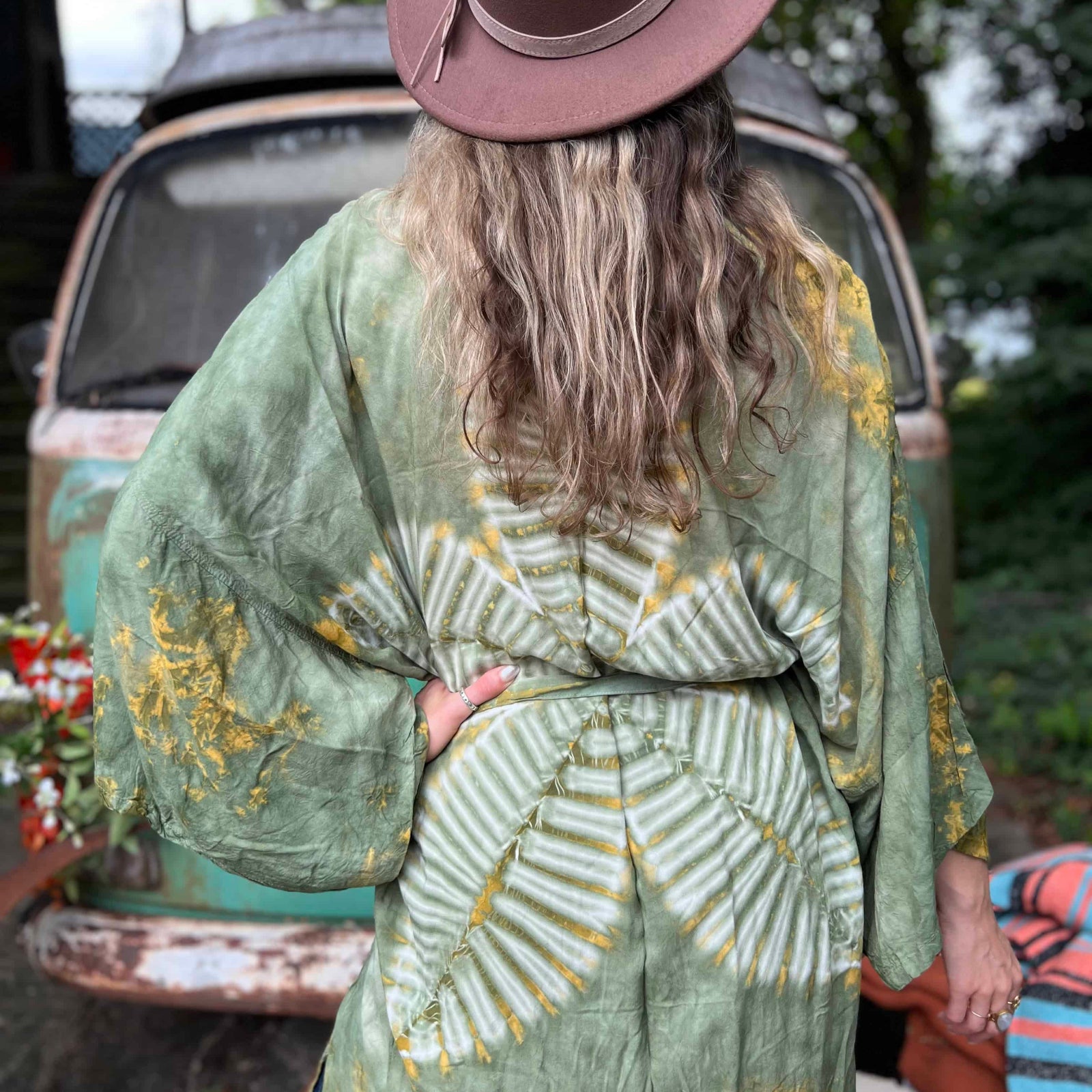 Person wearing a green patterned kimono and brown hat, standing outdoors with a vintage car in the background.