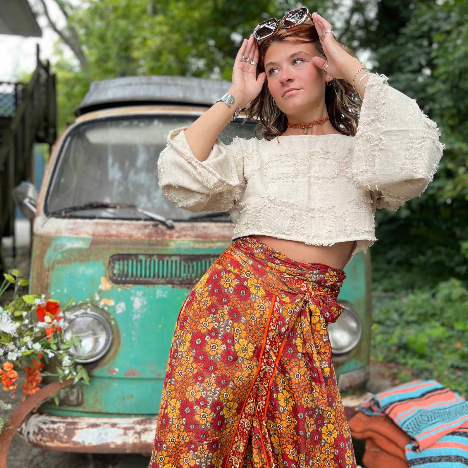 Woman in a white blouse and colorful skirt standing in front of a vintage car with greenery in the background