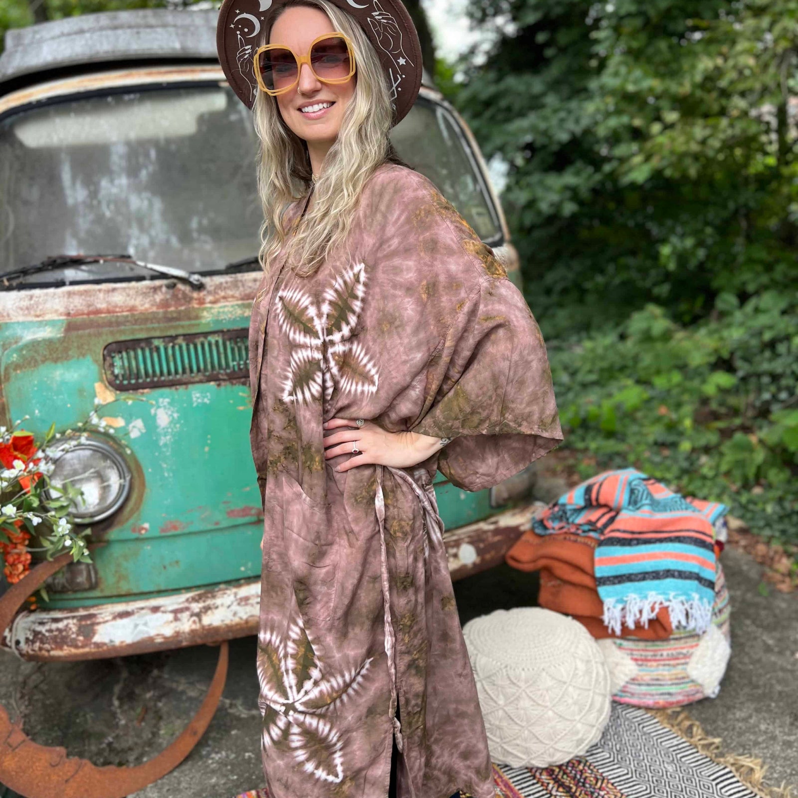 Woman in a patterned robe standing in front of a vintage car with a colorful rug and pillows on the ground.